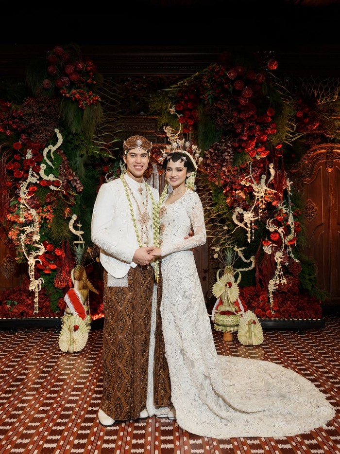 Di hari akad, Syifa tampil lebih sakral dalam kebaya putih panjang berbalut detail lace dan payet halus yang elegan. Riasan adat Jawa dengan paes, serta aksesori tradisional modern, memperkuat kesan anggun yang tidak berlebihan. Dipadukan dengan kain batik sidomukti, tampilannya memancarkan makna ketenangan dan harapan akan kehidupan rumah tangga yang sejahtera dan harmonis.