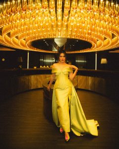 Gaun bergaya off-shoulder tersebut dipadukan dengan detail korset pada bagian bodi yang membentuk siluet tubuh secara elegan. Aksen ekor panjang yang menjuntai memberikan sentuhan glamor, sementara perhiasan minimalis, riasan bernuansa natural, dan tatanan rambut yang sederhana membuat keseluruhan penampilannya tetap terlihat berkelas tanpa terkesan berlebihan.